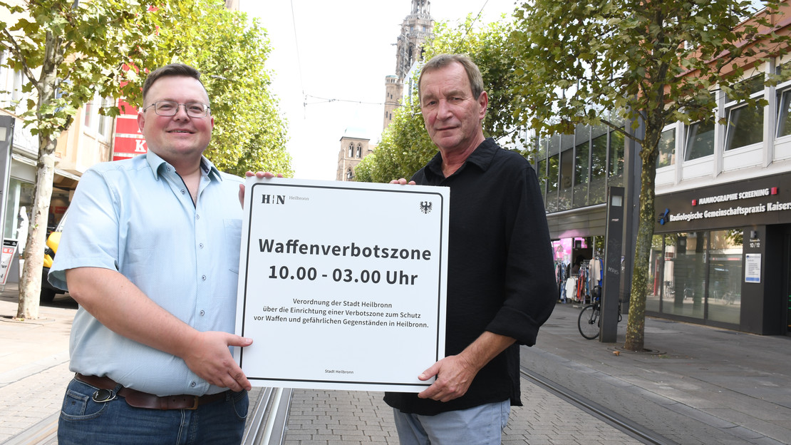 Das Bild zeigt zwei Ordnungsamtsmitarbeiter, die das neue Schild zur Waffenverbotszone Innenstadt in der Kaiserstraße präsentieren.