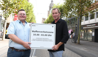 Das Bild zeigt zwei Ordnungsamtsmitarbeiter, die das neue Schild zur Waffenverbotszone Innenstadt in der Kaiserstraße präsentieren.