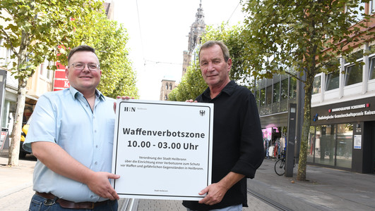 Das Bild zeigt zwei Ordnungsamtsmitarbeiter, die das neue Schild zur Waffenverbotszone Innenstadt in der Kaiserstraße präsentieren. 