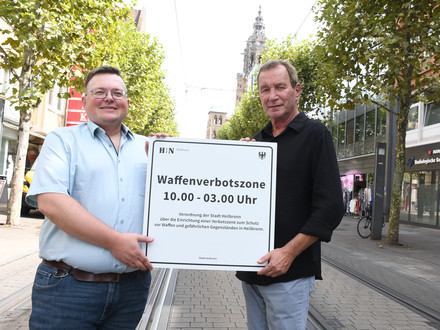 Das Bild zeigt zwei Ordnungsamtsmitarbeiter, die das neue Schild zur Waffenverbotszone Innenstadt in der Kaiserstraße präsentieren.