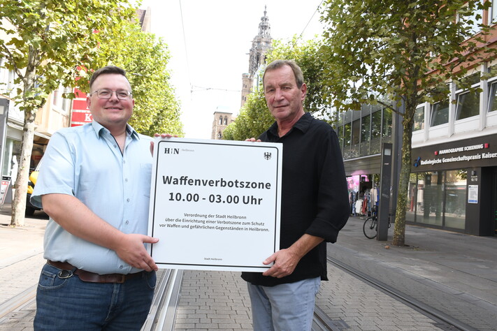 Das Bild zeigt zwei Ordnungsamtsmitarbeiter, die das neue Schild zur Waffenverbotszone Innenstadt in der Kaiserstraße präsentieren.