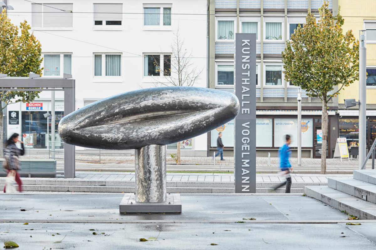 Skulptur "It's like a rock" von Richard Deacon, 2015 - Foto: Dietmar Strauß, Besigheim - Das Bild zeigt die Skulptur It’s like a rock von Richard Deacon aus dem Jahr 2015. Die glänzende, organisch geformte Skulptur erinnert an einen länglichen Stein oder ein abstrahiertes Objekt und ist auf einem Sockel befestigt. Sie steht auf einem offenen Platz im städtischen Raum. Im Hintergrund sind Gebäude, Bäume und vereinzelt vorbeigehende Menschen zu sehen. Die Skulptur wirkt modern und fügt sich als Kunstwerk in die urbane Umgebung ein.