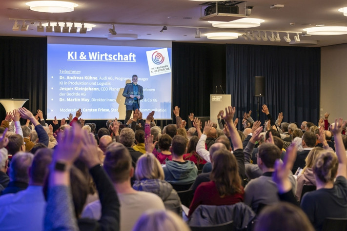 Viele Besucherinnen und Besucher sitzen bei einer Veranstaltung der Heilbronner KI-Gespräche in einem Vortragssaal und melden sich während einer Diskussion zu Wort. Auf der Bühne ist eine Präsentation zum Thema „KI & Wirtschaft“ zu sehen.
