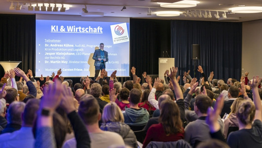 Viele Besucherinnen und Besucher sitzen bei einer Veranstaltung der Heilbronner KI-Gespräche in einem Vortragssaal und melden sich während einer Diskussion zu Wort. Auf der Bühne ist eine Präsentation zum Thema „KI & Wirtschaft“ zu sehen.