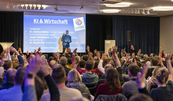 Viele Besucherinnen und Besucher sitzen bei einer Veranstaltung der Heilbronner KI-Gespräche in einem Vortragssaal und melden sich während einer Diskussion zu Wort. Auf der Bühne ist eine Präsentation zum Thema „KI & Wirtschaft“ zu sehen.