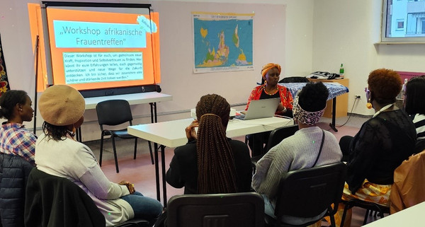 Workshop des afrikanische Frauentreffs mit mehreren Teilnehmerinnen die an Tischen sitzen und einer Referentin zuhören die eine Präsentation zeigt