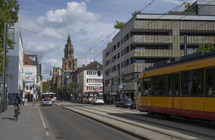 Das Bild zeigt den gleichen Stadtausschnitt im Sommer 2026. Moderne , lange Stadtbahnen sind zu sehen, die leicht veränderte Kilianskirche und als Ankerpunkt das Marra-Haus in der rechten Bildhälfte. 