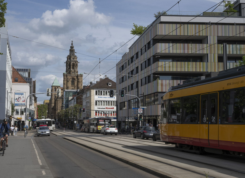 Das Bild zeigt den gleichen Stadtausschnitt im Sommer 2026. Moderne , lange Stadtbahnen sind zu sehen, die leicht veränderte Kilianskirche und als Ankerpunkt das Marra-Haus in der rechten Bildhälfte. 