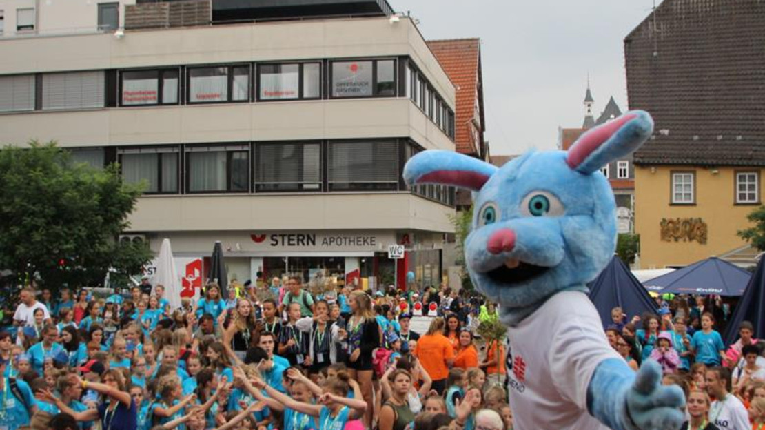 Das Bild zeigt eine große Schar fröhlicher Kinder auf einem Platz, vorne steht ein Maskottchen in Sportdress. 