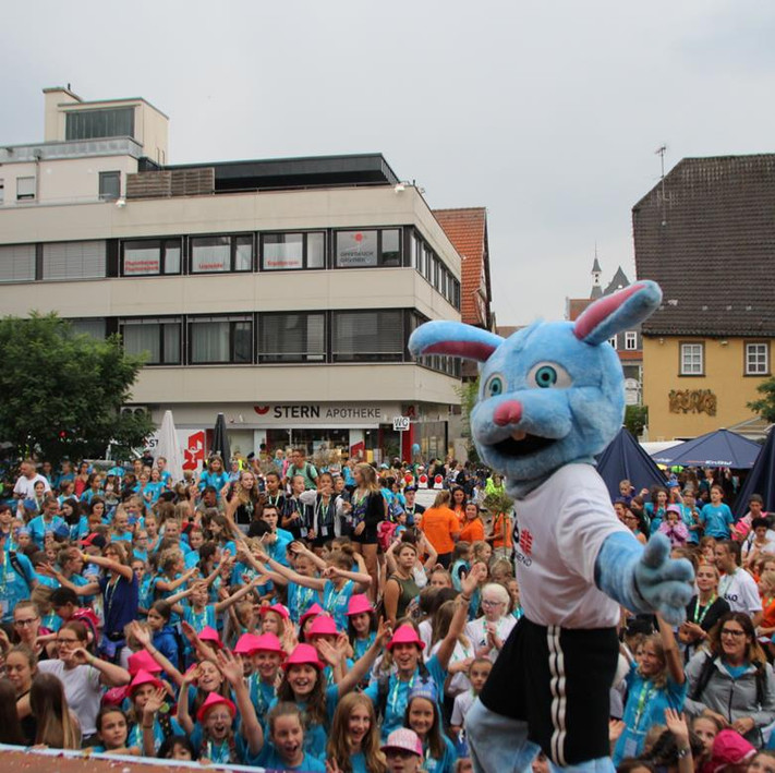 Das Bild zeigt eine große Schar fröhlicher Kinder auf einem Platz, vorne steht ein Maskottchen in Sportdress. 