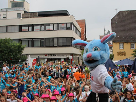 Das Bild zeigt eine große Schar fröhlicher Kinder auf einem Platz, vorne steht ein Maskottchen in Sportdress. 