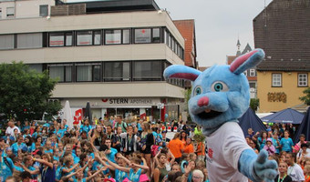 Das Bild zeigt eine große Schar fröhlicher Kinder auf einem Platz, vorne steht ein Maskottchen in Sportdress.