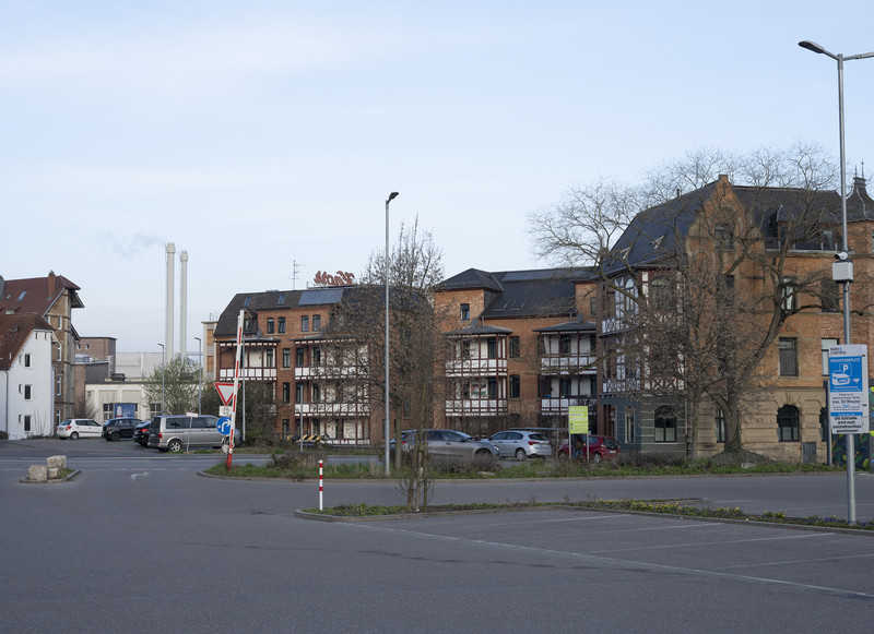 Das Bild zeigt einen großen Parkplatz in Vordergrund, der zu einem großen Lebensmittelmarkt gehört, der auf dem Areal des früheren Südbahnhofs entstand. Die Firma Knorr hat hier noch ihren Sitz. Die Wohngebäude sind erhalten geblieben und modernisiert worden. Autos haben verstärkt den öffentlichen Raum erobert. 