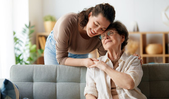 Das Bild zeigt eine junge Betreuerin, die eine Seniorin in deren Wohnung in dem Arm nimmt.