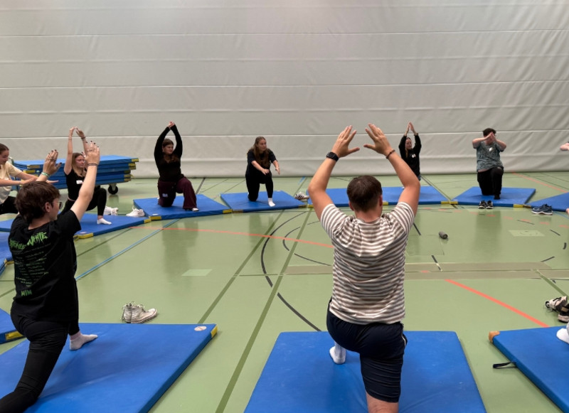 Auszubildende und Studierende absolvieren gemeinsam Übungen auf Matten in einer Sporthalle.