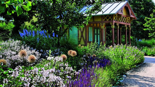 Das Bild zeigt bunt blühende Blumen und eine historische Gartenlaube im Hintergrund. 