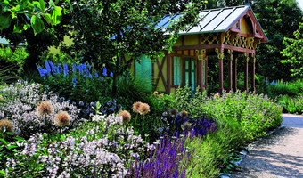 Das Bild zeigt bunt blühende Blumen und eine historische Gartenlaube im Hintergrund. 
