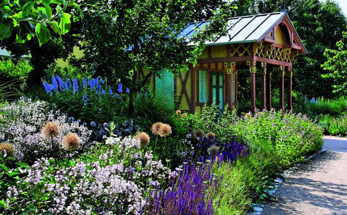 Das Bild zeigt bunt blühende Blumen und eine historische Gartenlaube im Hintergrund.