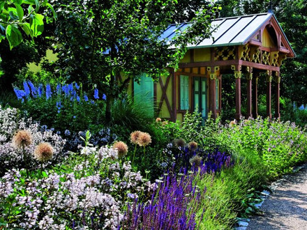 Das Bild zeigt bunt blühende Blumen und eine historische Gartenlaube im Hintergrund. 