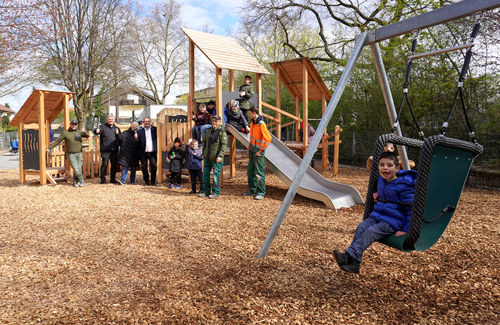 Ein Spielplatz auf dem einige Erwachsene stehen und Kinder spielen. Die Spielgeräte sind aus Holz.