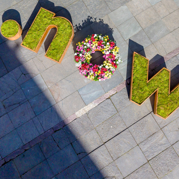 Begrünter Schriftzug ".now" aus Moos und Blumen auf gepflastertem Platz aus der Vogelperspektive