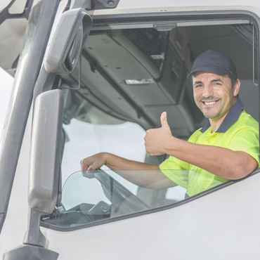 Ein lächelnder Lkw Fahrer sitzt in der Fahrerkabine und zeigt den Daumen nach oben. Er trägt eine gelbe Warnweste und eine dunkle Kappe.