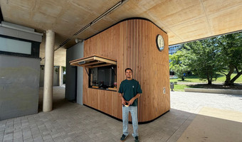 Auf dem Foto ist ein junger Mann vor einem Schulkiosk zu sehen. Er hält eine Brezel in der Hand. Der Kiosk aus Holz hinter ihm wurde von ihm entworfen.