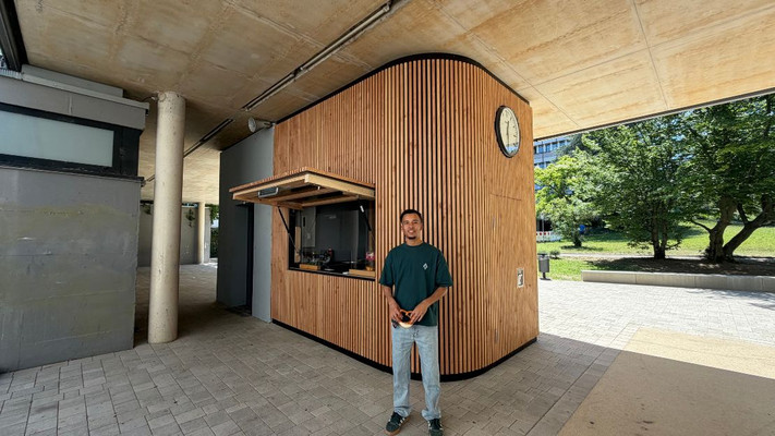 Auf dem Foto ist ein junger Mann vor einem Schulkiosk zu sehen. Er hält eine Brezel in der Hand. Der Kiosk aus Holz hinter ihm wurde von ihm entworfen.