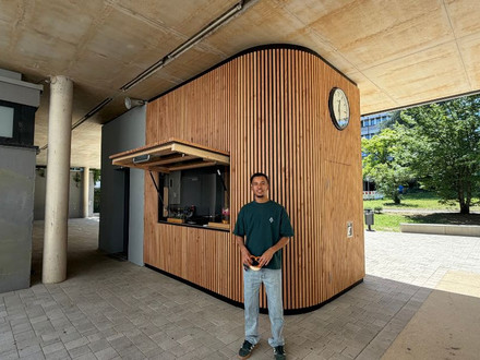 Auf dem Foto ist ein junger Mann vor einem Schulkiosk zu sehen. Er hält eine Brezel in der Hand. Der Kiosk aus Holz hinter ihm wurde von ihm entworfen. 
