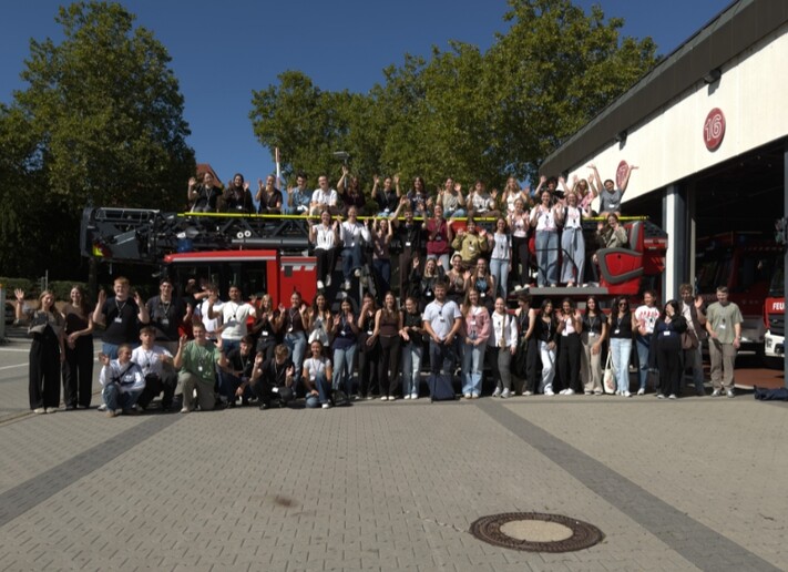 Die neuen Auszubildenden und Studierenden stehen gemeinsam mit Einsatzfahrzeugen vor der Feuerwache und winken in die Kamera.