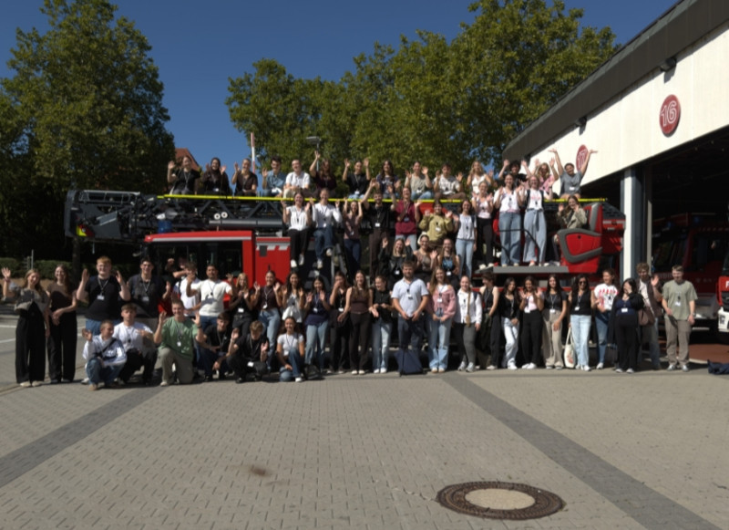 Die neuen Auszubildenden und Studierenden stehen gemeinsam mit Einsatzfahrzeugen vor der Feuerwache und winken in die Kamera.