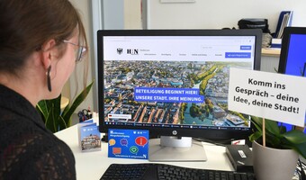 Eine Frau sitzt an einem Schreibtisch und betrachtet auf einem Computerbildschirm die Website der Heilbronner Bürgerbeteiligung. Neben dem Monitor steht ein Schild mit der Aufschrift „Komm ins Gespräch – deine Idee, deine Stadt!“.