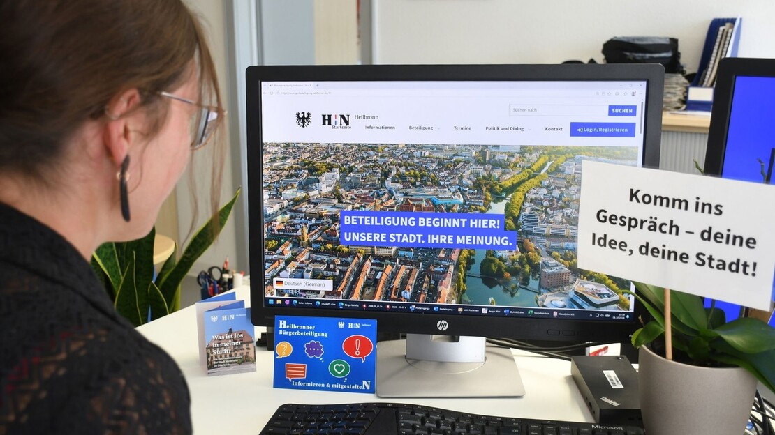 Eine Frau sitzt an einem Schreibtisch und betrachtet auf einem Computerbildschirm die Website der Heilbronner Bürgerbeteiligung. Neben dem Monitor steht ein Schild mit der Aufschrift „Komm ins Gespräch – deine Idee, deine Stadt!“.