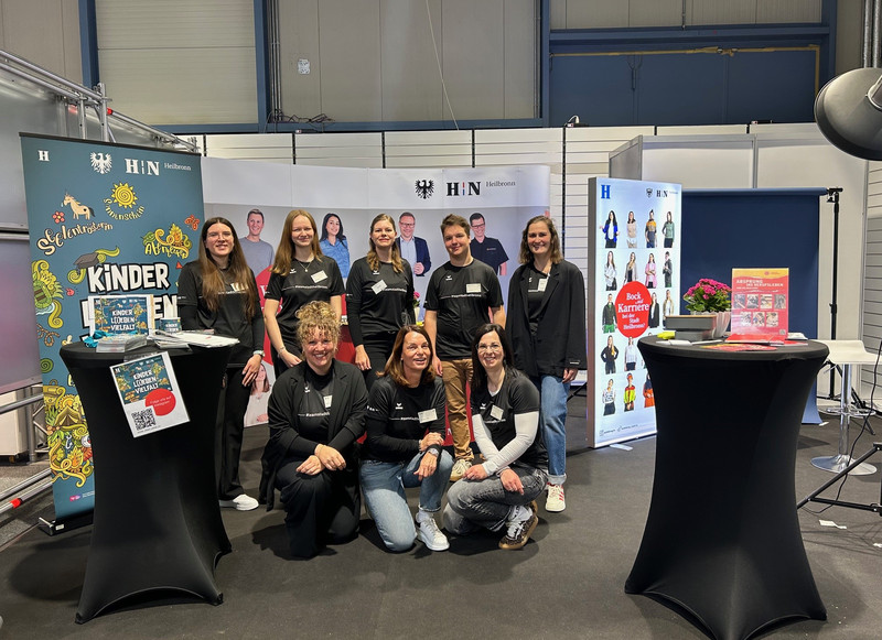 Das Bild zeigt eine Gruppe Auszubildender und Betreuender der Stadtverwaltung an einem Infostand mit vielen Plakaten auf der Bildungsmesse. 