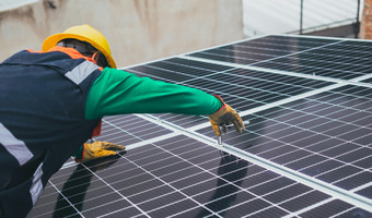 Ein Mann mit einem Bauarbeiterhelm baut auf einem Dach eine Solaranlage, Photovoltaikanlage an.