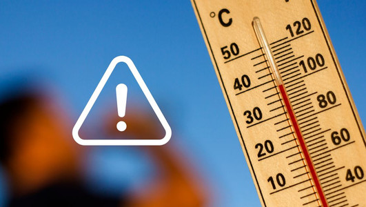 Nahaufnahme eines Thermometers mit hoher Temperaturanzeige vor blauem Himmel. Ein weißes Warnsymbol mit Ausrufezeichen weist auf die gesundheitlichen Risiken großer Hitze hin.