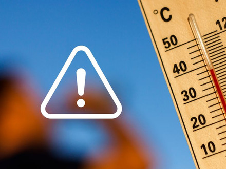 Nahaufnahme eines Thermometers mit hoher Temperaturanzeige vor blauem Himmel. Ein weißes Warnsymbol mit Ausrufezeichen weist auf die gesundheitlichen Risiken großer Hitze hin.