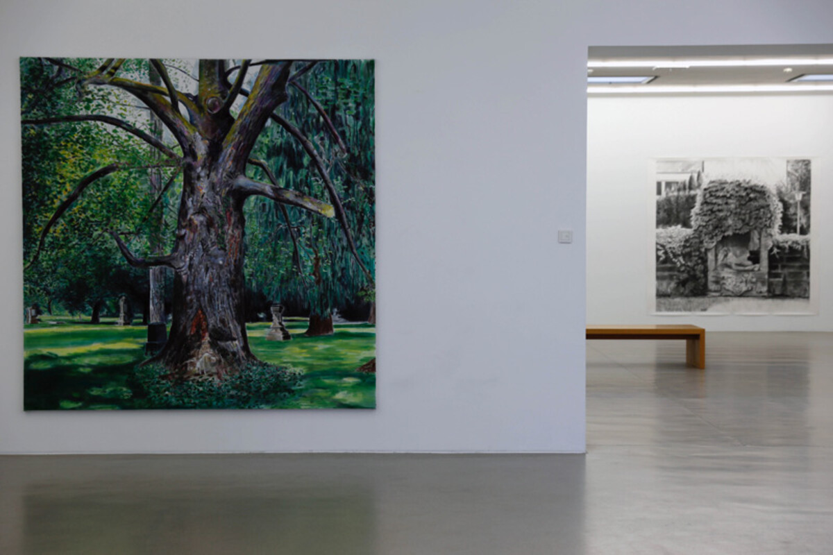 Das Bild zeigt eine Ausstellungssituation im Kunstverein Heilbronn. An einer weißen Wand hängt links ein großformatiges Gemälde eines Baumes in einer grünen Landschaft. Rechts öffnet sich der Blick in einen weiteren Raum, in dem zusätzliche Kunstwerke an den Wänden präsentiert werden. Der Ausstellungsraum ist hell, minimalistisch und ruhig gestaltet. Die Arbeiten stammen von Yves Belorgey und waren Teil einer Ausstellung im Jahr 2019. Yves Belorgey, Ausstellungsansicht Kunstverein Heilbronn 2019 - Foto: Yves Belorgey