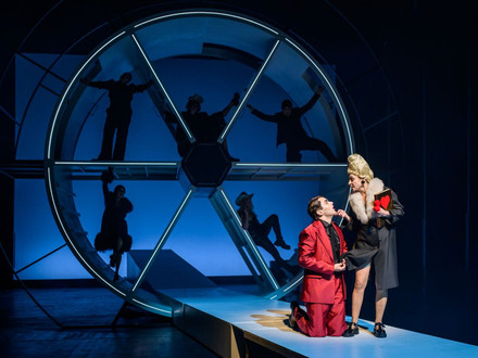 Das Bild zeigt eine Schauspielerin und einen Schauspieler vor einem Bühnenbild mit Schattenelementen. 