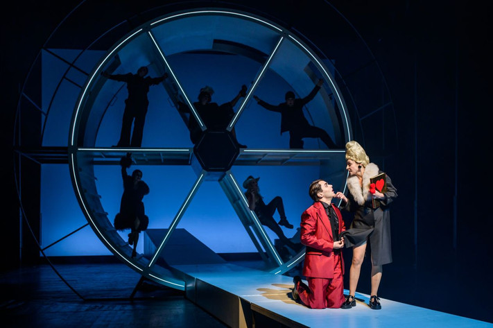 Das Bild zeigt eine Schauspielerin und einen Schauspieler vor einem Bühnenbild mit Schattenelementen. 