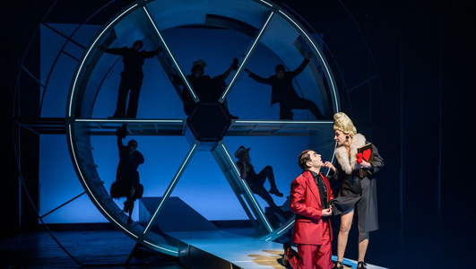 Das Bild zeigt eine Schauspielerin und einen Schauspieler vor einem Bühnenbild mit Schattenelementen. 