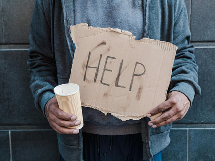 Das Bild zeigt einen Menschen in einfacher Kleidung, der ein Schild mit dem Wort "Help" für "Hilfe in den Händen hält. 