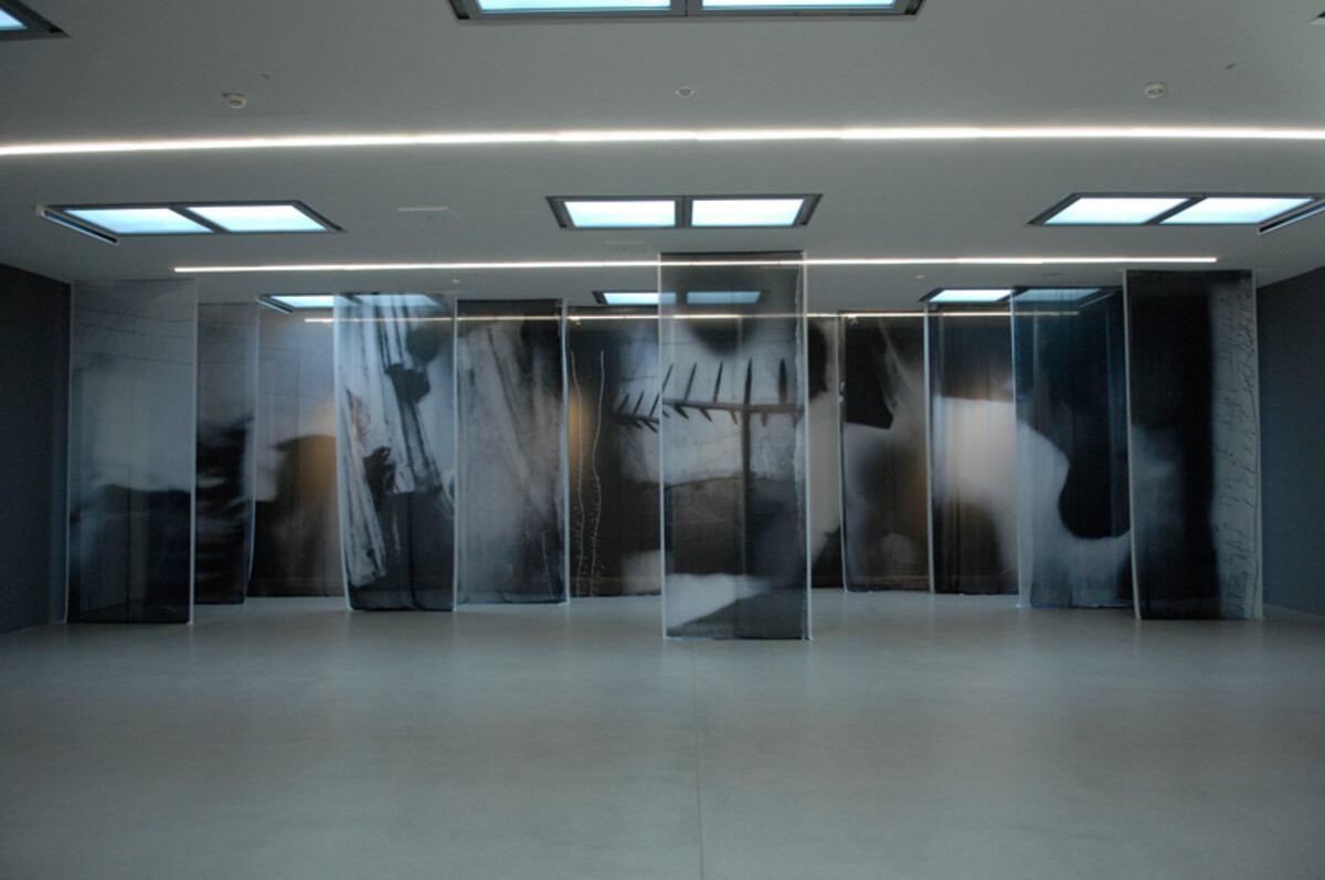 Das Bild zeigt eine Ausstellungssituation im Kunstverein Heilbronn. In einem großen, hellen Raum stehen mehrere hohe, halbtransparente Bildtafeln in einer Reihe. Auf ihnen sind abstrakte, fotografisch wirkende Motive in dunklen und hellen Flächen zu sehen. Die Tafeln sind so angeordnet, dass Besucherinnen und Besucher zwischen ihnen hindurchgehen können. Die Installation stammt von Sergej Vutuc und war Teil einer Ausstellung im Jahr 2019. Sergej Vutuc, Ausstellungsansicht Kunstverein Heilbronn 2019 - Foto: Sergej Vutuc