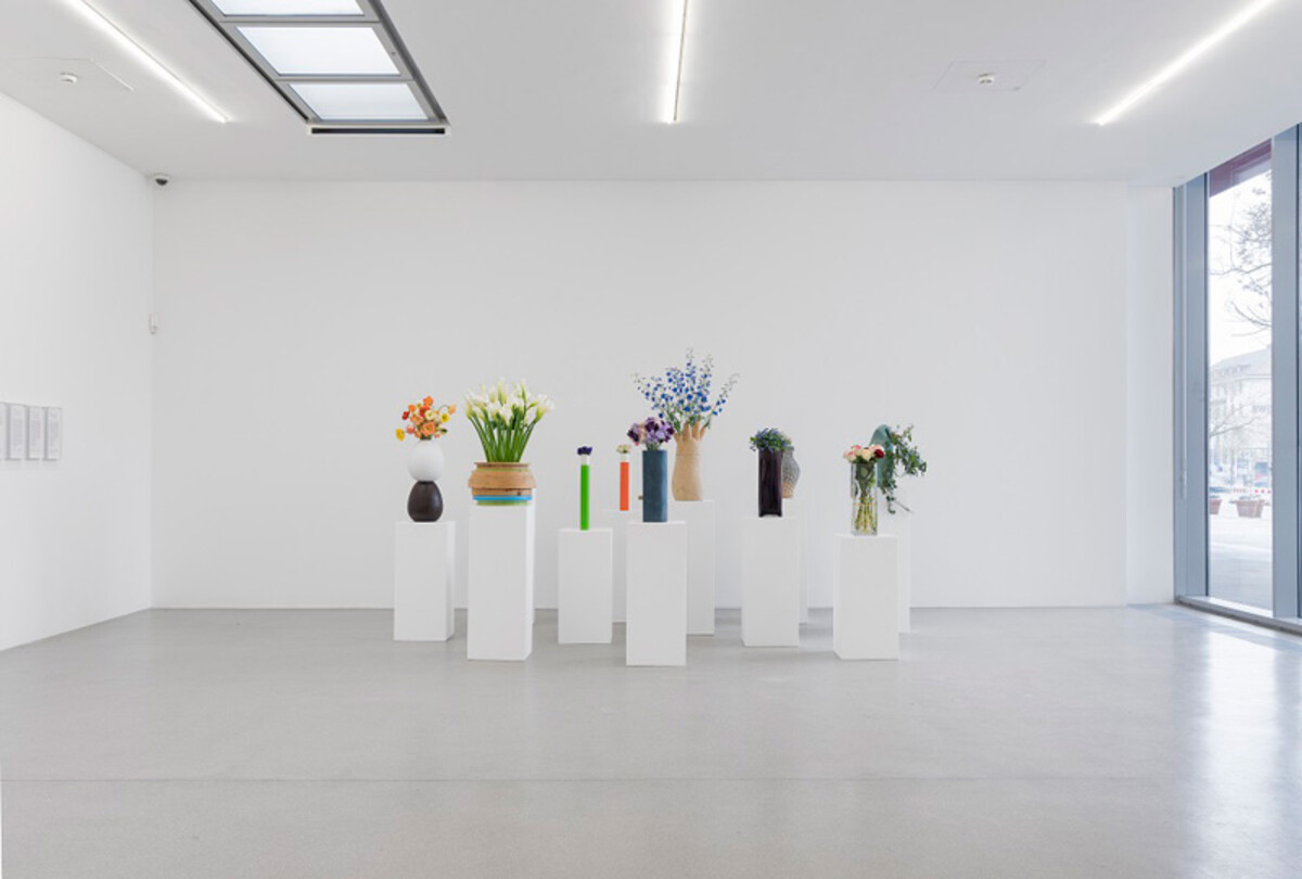 Das Bild zeigt eine minimalistisch gestaltete Ausstellungsszene im Kunstverein Heilbronn. In einem hellen, nahezu leeren Raum stehen mehrere weiße Sockel in einer Reihe. Auf jedem Sockel befindet sich eine Vase mit unterschiedlich arrangierten Blumen. Die Installation trägt den Titel Familienporträt, Durch die Blume von Tobias Rehberger aus dem Jahr 2019. Die klare Präsentation lenkt den Fokus auf die individuellen Blumenarrangements und deren symbolische Wirkung. Tobias Rehberger, Familienporträt, Durch die Blume, Kuntsverein Heilbronn 2019 - Foto: Wolfgang Günzel, Frankfurt