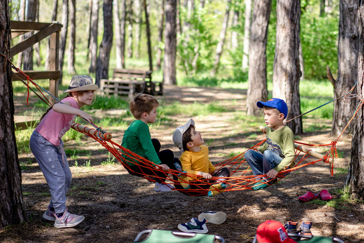 Das Bild zeigt spielende Kinder in einer Hängematte zwischen Bäumen. Es thematisiert das breite Angebot des Sommerferienprogramms. 