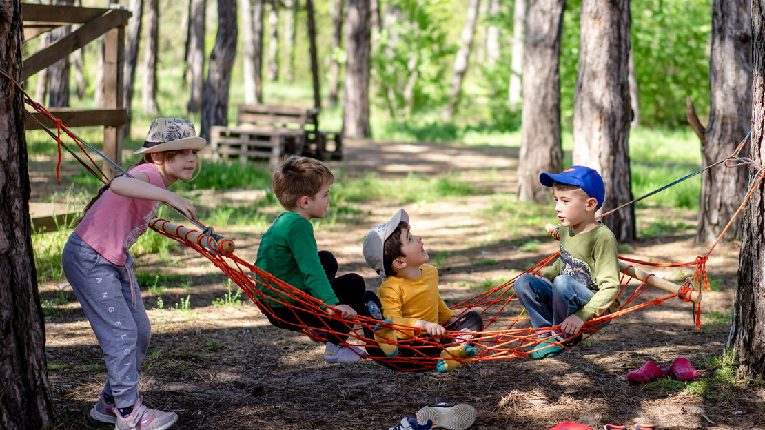 Das Bild zeigt spielende Kinder in einer Hängematte zwischen Bäumen. Es thematisiert das breite Angebot des Sommerferienprogramms.