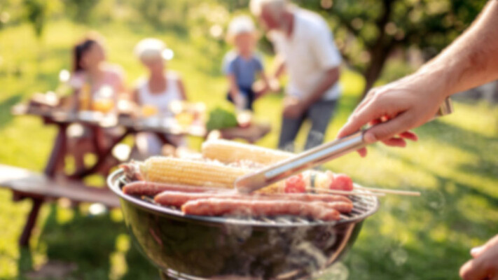 Grill mit Würstchen und Maiskolben, im Hintergrund vier Personen