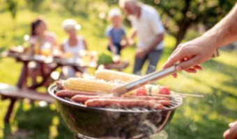 Grill mit Würstchen und Maiskolben, im Hintergrund vier Personen