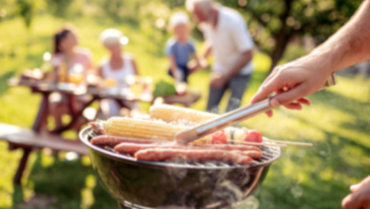 Grill mit Würstchen und Maiskolben, im Hintergrund vier Personen