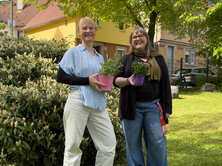 Tina Wenk links und Johanna Greef stehen im Freien vor einem Wohngebäude und halten jeweils einen Blumentopf mit Pflanzen in den Händen. Beide lächeln in die Kamera, im Hintergrund sind Sträucher und ein Baum zu sehen.